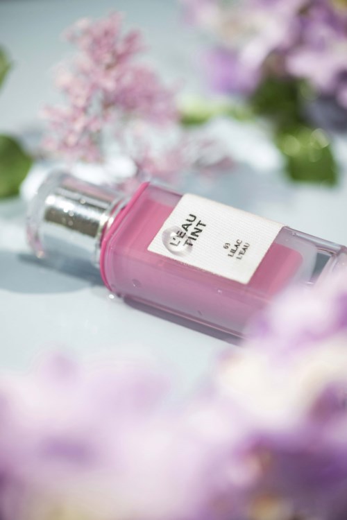 Een roze parfumfles met de tekst "Eau Tain" ligt tussen roze en paarse bloemen.