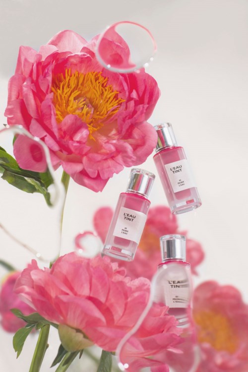 Drie kleine roze parfumflesjes hangen tussen roze pioenen met waterdruppels, wat een zachte bloemrijke scène creëert.