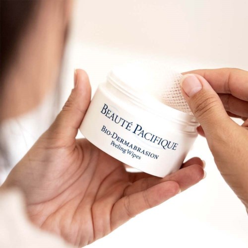 En person håller i en vit burk med etiketten 'BEAUTE PACIFIQUE' och avskrapningsservetter inside.