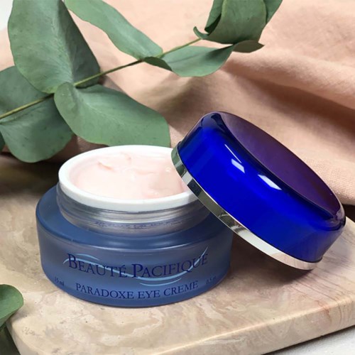Ein blauer Behälter mit Augencreme, offener Deckel, grüne Blätter und beiger Hintergrund. Beschriftung: 'BEAUTE PACIFIQUE'.