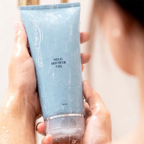 Henkilö pitää sinistä suihkugeeliä, jossa lukee 'Mild Shower Gel'. Käsissä on vettä.