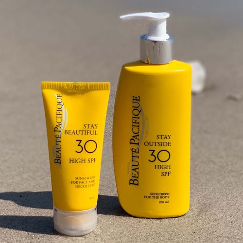 Zwei gelbe Sonnencremeflaschen mit schwarzem Text, eine klein und eine groß, am Strand mit Sand und Schatten.