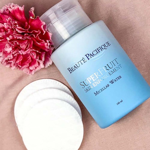 A bottle of micellar water with cotton pads and a pink flower beside it on a soft surface.