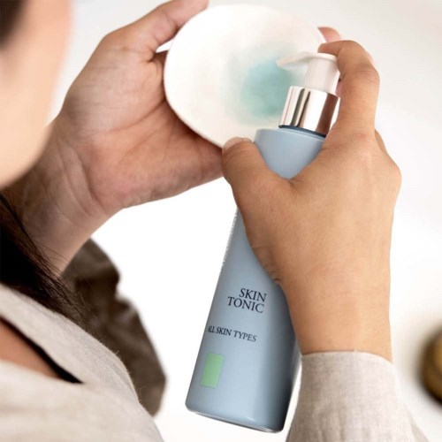 A person uses a blue bottle labeled 'Skin Tonic' to apply a liquid onto a cotton pad.