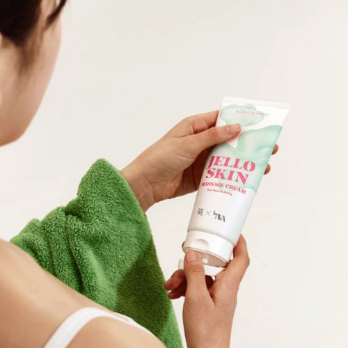 En person håller ett rör med Hello Skin massagekräm mot en enkel bakgrund. De har en grön handduk på axeln.