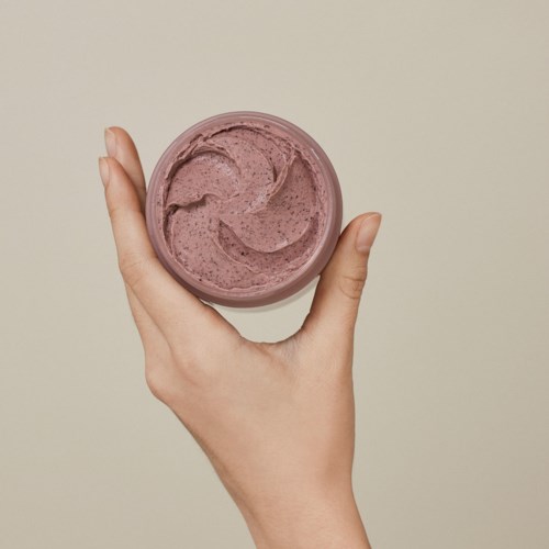 Eine Hand hält einen kleinen runden Behälter mit lila Creme oder Gel vor einem schlichten Hintergrund.