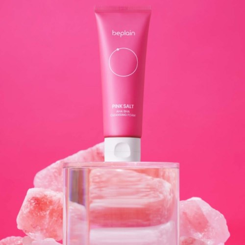 Een roze tube van Beplain Pink Salt reiningsschuim boven een glas met roze zoutkristallen op een roze achtergrond.