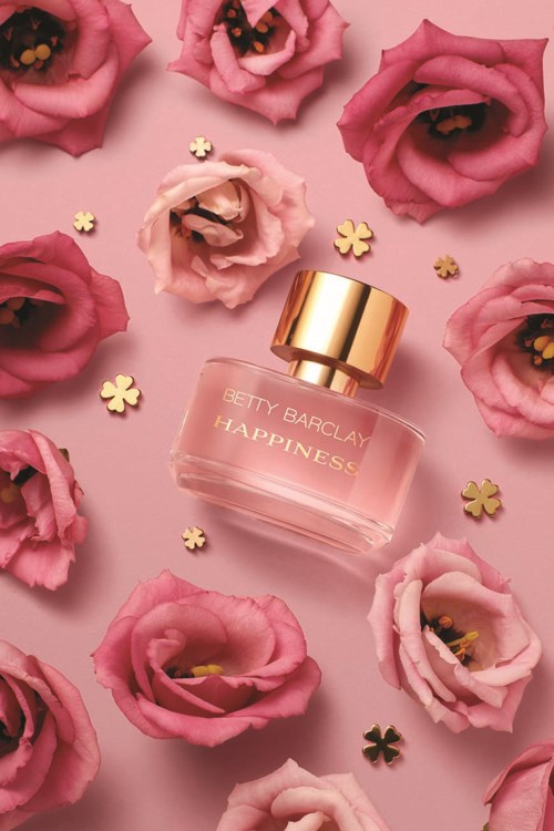 Een parfumfles genaamd Betty Barclay Happiness, omgeven door roze en rode bloemen en gouden bloemenconfetti.