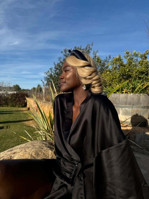 A woman with blonde hair wears a black robe, sits outdoors with green plants and blue sky behind her.