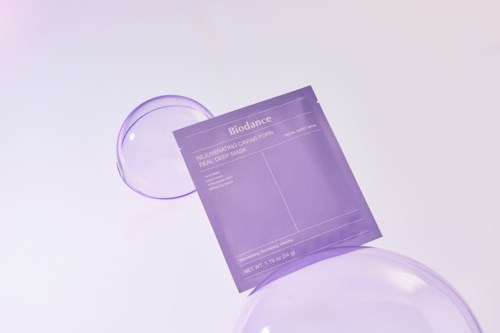 A purple skincare product packet with a transparent face mask, surrounded by clear glass spheres on a light background.