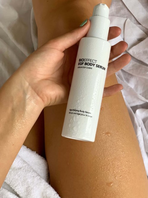 A person holds a white bottle labeled 'Bioeffect EGF Body Serum' with water droplets on their skin.