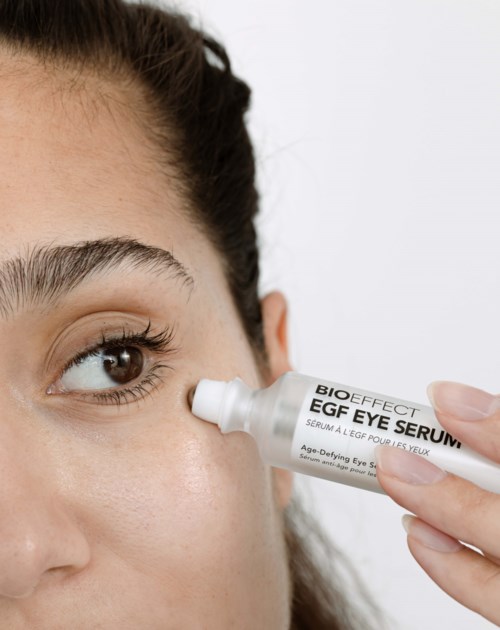A woman applies eye serum under her eye with a white dropper bottle.