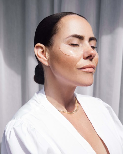 A woman with smooth skin, closed eyes, and dark hair in a bun, wearing a white top and a gold necklace.