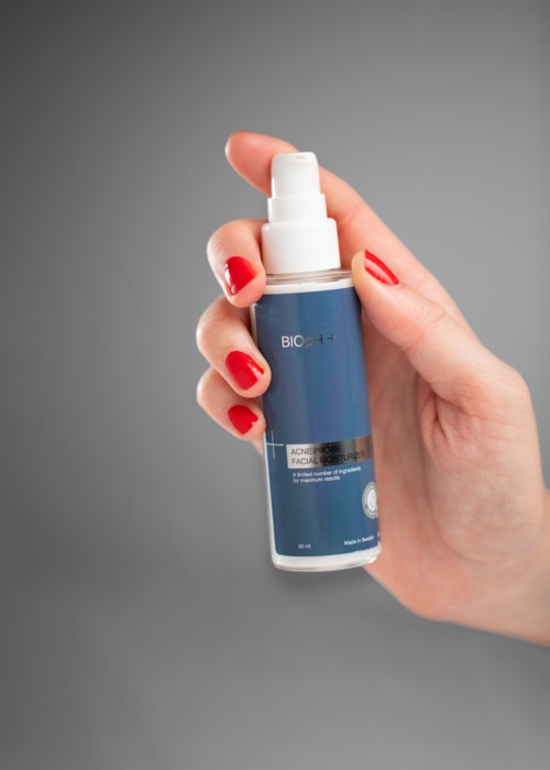 En hand med röda naglar håller en blå och vit sprayflaska mot en grå bakgrund.