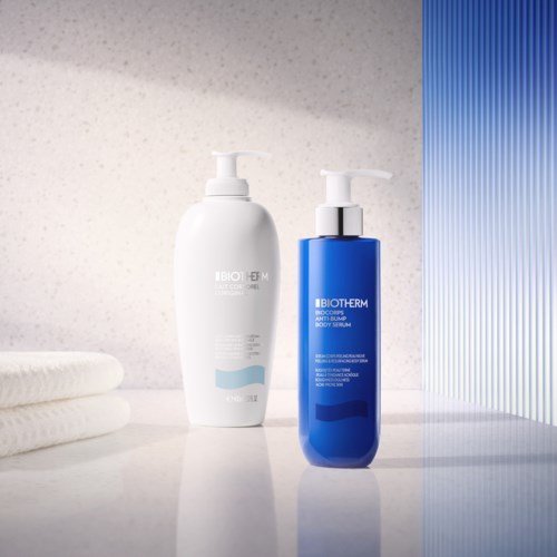 Zwei Flaschen Hautpflegeprodukte auf einer weißen Oberfläche, eine weiß und eine blau, mit gefalteten Handtüchern in der Nähe.