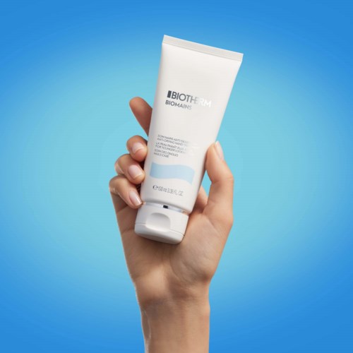 Een hand houdt een witte tube huidverzorgingsproduct vast tegen een blauwe achtergrond.