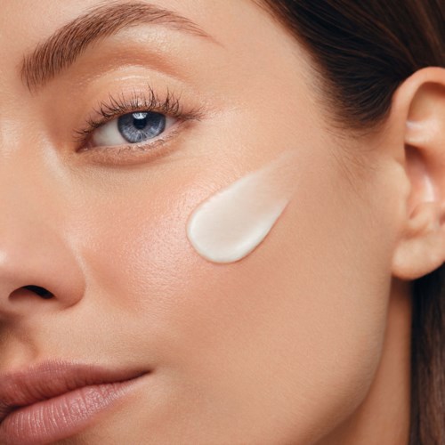 Een vrouw met blauwe ogen heeft een crème op haar wang aangebracht, toont een egale huid en natuurlijke make-up.