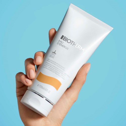 En hand håller i ett vitt rör märkt 'BIOTHERM Eau D'Energie' mot en blå bakgrund.