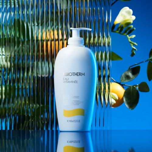 A white bottle labeled 'Biotherm Eau Vitaminee' against a colorful, textured background with green leaves and white flowers.
