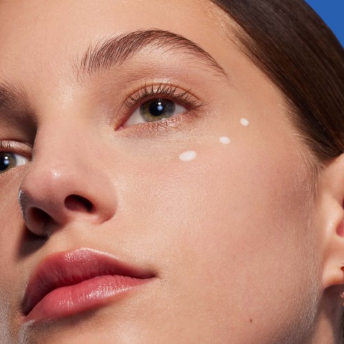 Een vrouw met lichte huid en blauwe ogen heeft witte druppels van crème of lotion op haar wang, vlakbij haar oog.