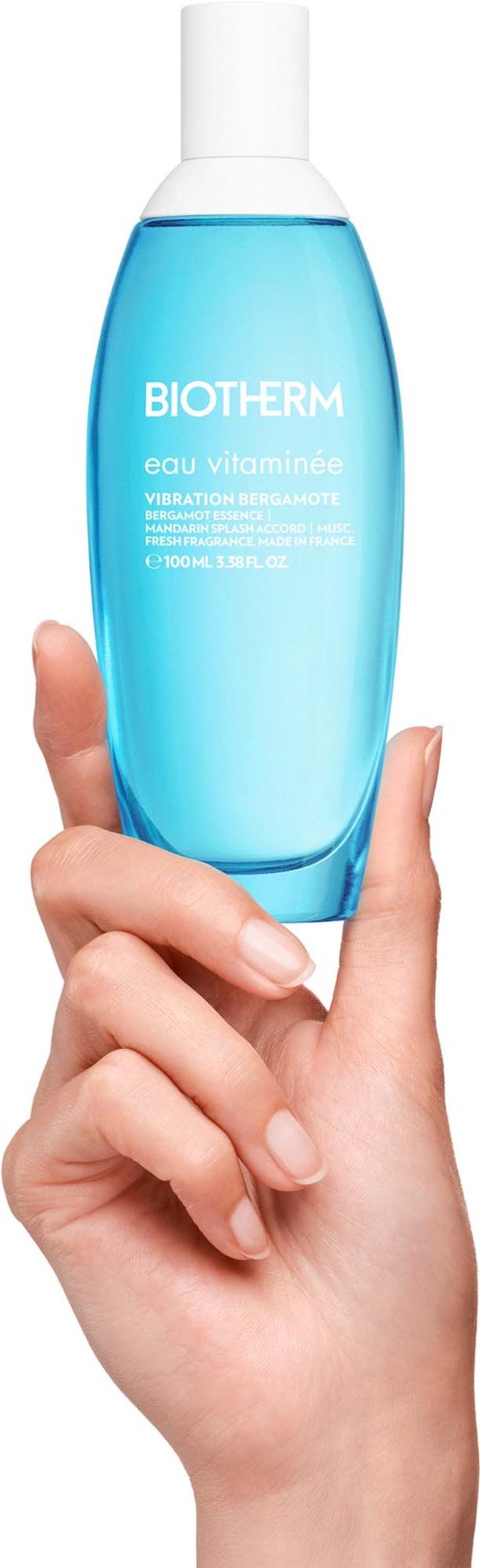 Een hand houdt een blauwe fles Biotherm Eau Vitaminée, een hydraterend huidproduct, tegen een witte achtergrond.