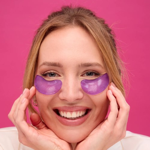 A woman smiles and wears purple under-eye patches against a pink background.