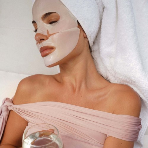 Kvinna med ansiktsmask, handduk runt huvudet, bär en rosa top, avkopplande på spa eller salong.