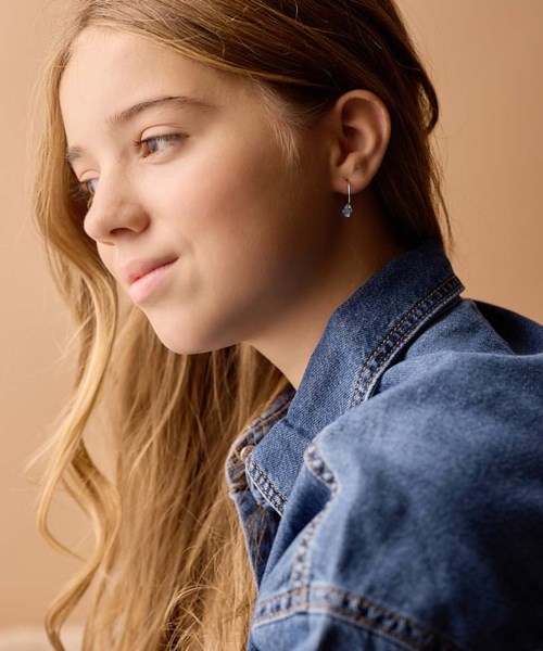 A young girl with long, wavy blonde hair wears a denim jacket and a small earring, looking sideways.