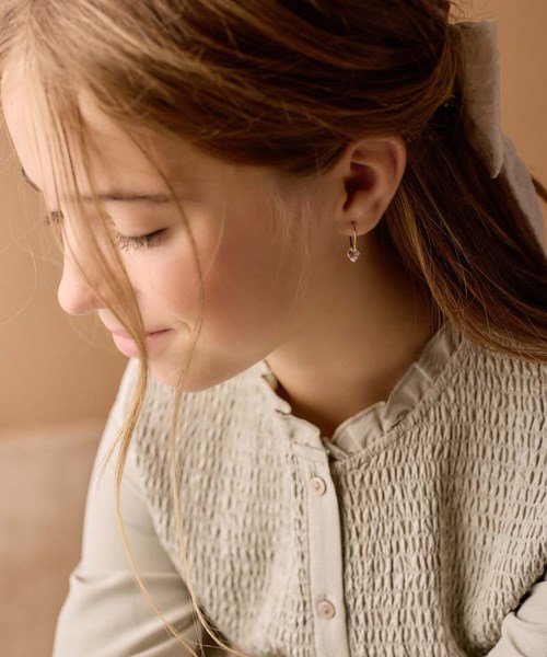 A girl with closed eyes, smiling, with long brown hair in a bun, wearing earrings and a cream blouse.