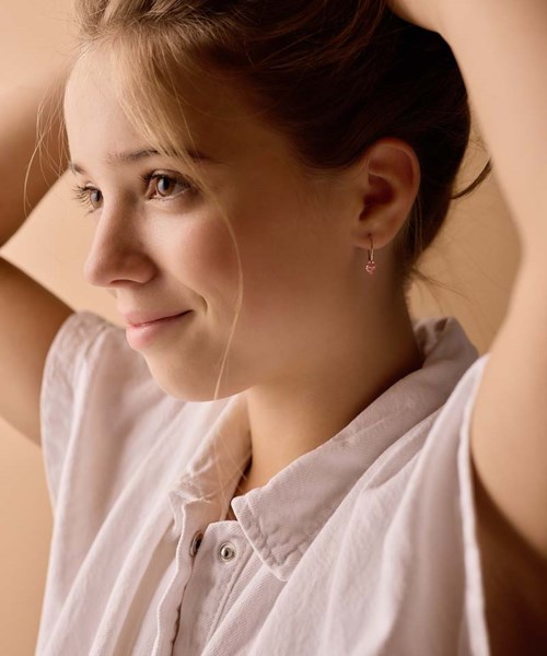 A young woman smiles softly, with her hands raised behind her head, wearing a white shirt and small earrings.