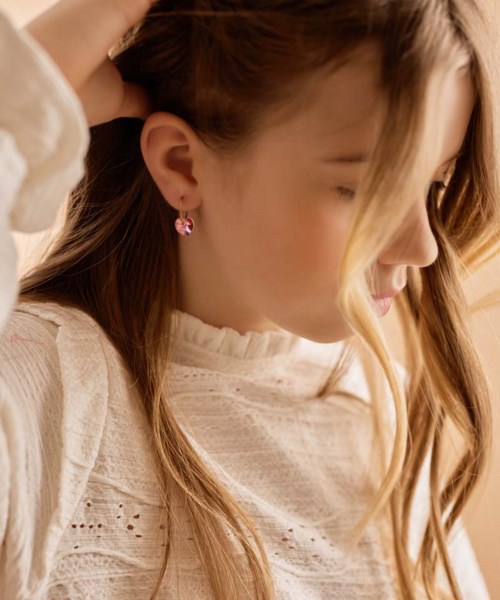 A young woman with long hair is smiling, wearing earrings and a white, patterned top, touching her hair gently.