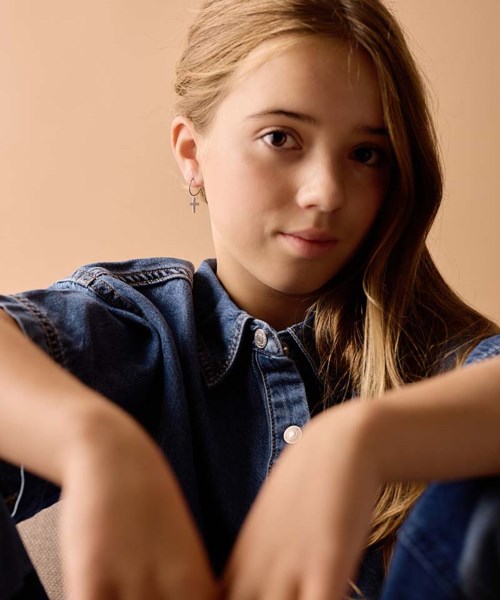 A girl with long hair wearing a denim shirt, sitting indoors, looking at the camera with a gentle expression.