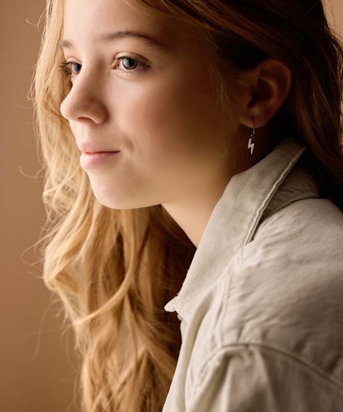 A young woman with long, wavy hair wears a lightning bolt earring, looking thoughtful against a plain background.