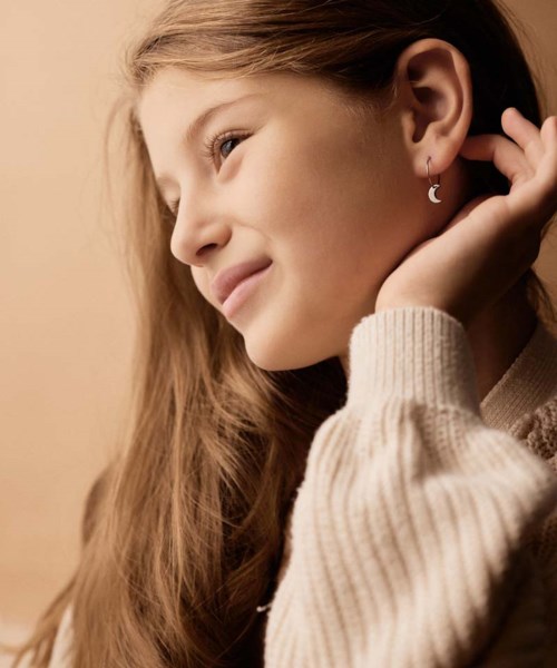 A girl with long hair wearing a sweater, touching her ear with a moon-shaped earring, smiling softly.