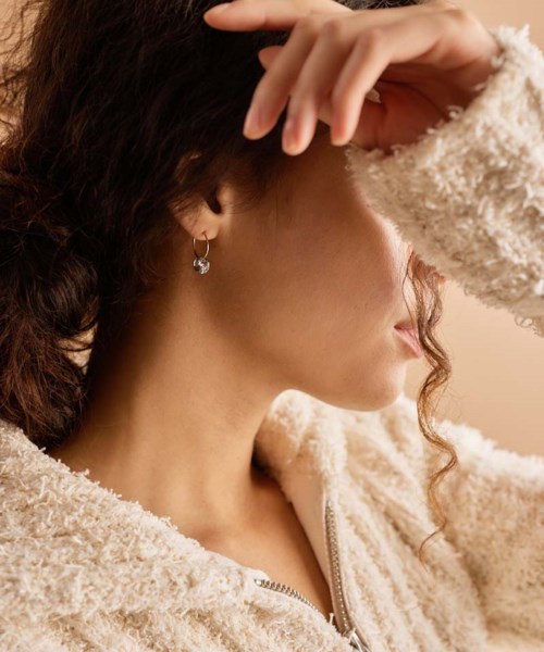 A woman wearing a fluffy beige jacket and earrings, with her hand partly covering her face, looking away.