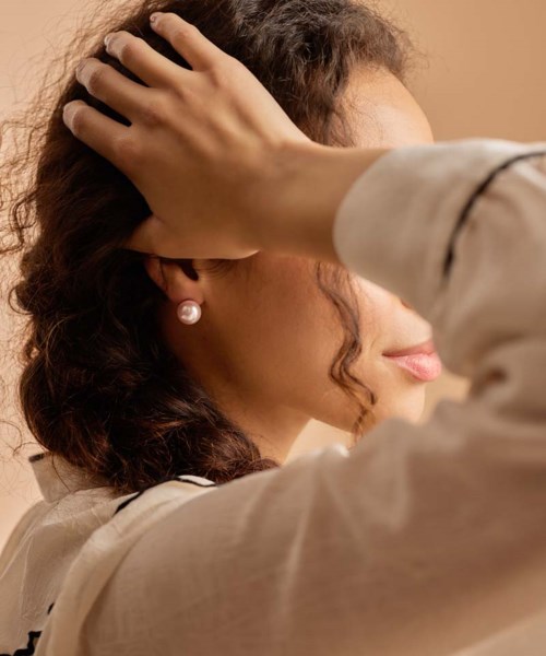 A woman with curly hair wears pearls, touching her head gently, with a neutral background.