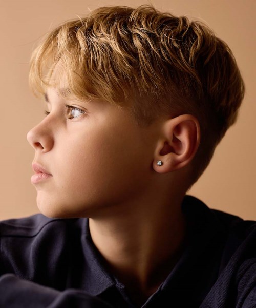 A young boy with blonde hair looking to the left, wearing a dark shirt and an earring, against a plain background.