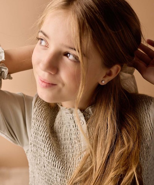 A young girl with blond hair and a gentle smile, looking upwards with her hand behind her head.