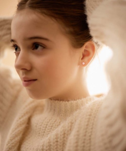Ein junges Mädchen mit braunen Haaren, trägt einen cremefarbenen Pullover und schaut seitlich mit einem sanften Lächeln.