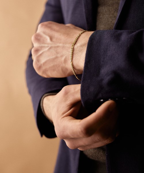 En person bär en mörk blazer med ett guldarmband, håller armen mot bröstet. Bakgrunden är enkel.