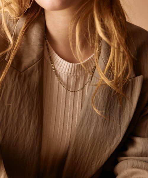 En kvinna i beige jacka och vit topp med layerade halsband, med långt, vågigt blont hår.