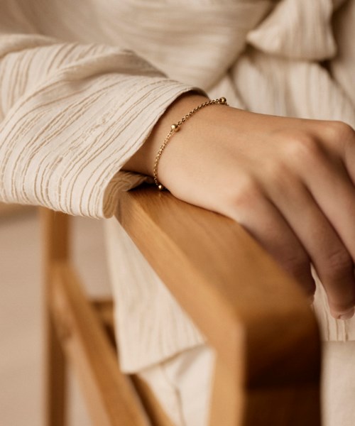 A person wears a gold bracelet, resting their hand on a wooden surface. They are dressed in a cream-colored, long-sleeved shirt.
