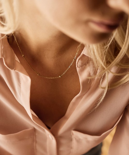 A woman wears a light pink shirt and a small gold necklace, with her face partly visible and blonde hair.