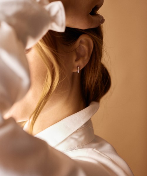 A woman with blonde hair and a single earring, dressed in a white shirt, is touching her head.