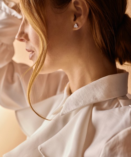 En kvinne med rødt hår og diamant-øreringer, iført en hvit skjorte, smiler forsiktig, med varmt lys og mykt bakgrunn.