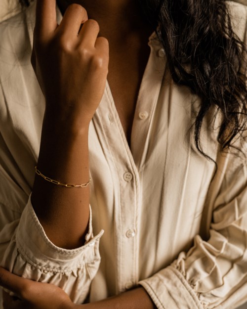A woman with dark, curly hair wearing a beige blouse, holding her hand near her face, wearing a bracelet.