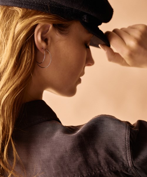 A woman with long hair, wearing a black hat and jacket, is touching her hat while looking down.