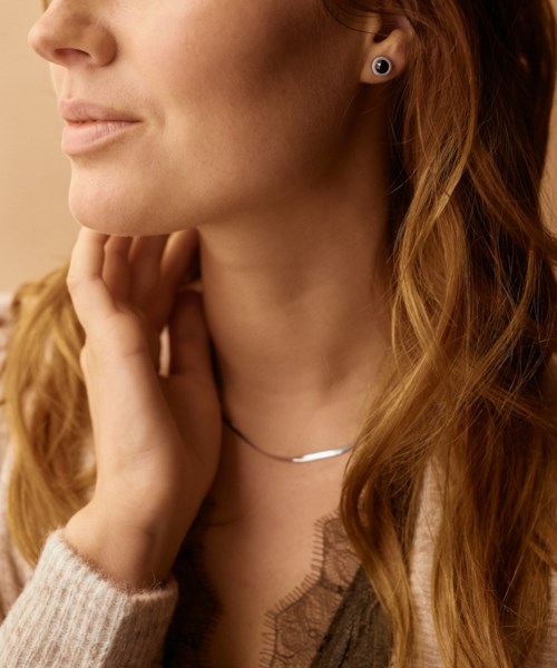 A woman with earrings and a necklace, touching her neck with her hand, wearing a beige sweater.