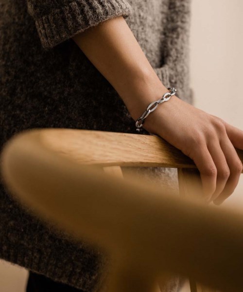 A person's hand with a silver chain bracelet rests on a wooden surface, wearing a cozy sweater.
