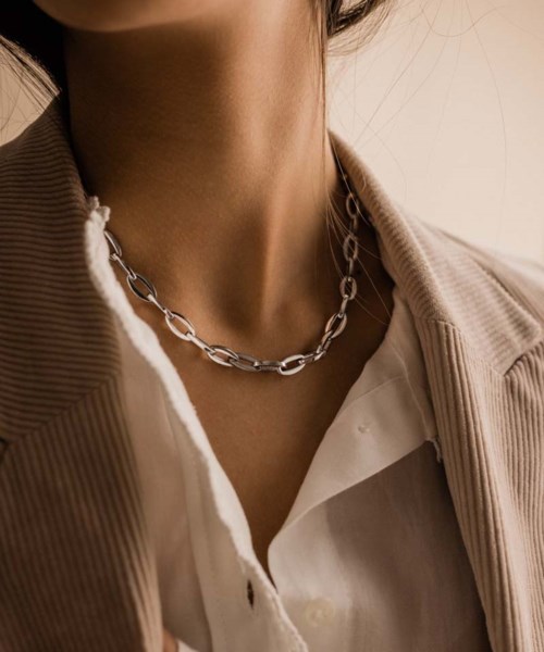 A person wears a chunky chain necklace over a white shirt and beige jacket, with a close-up focus on the neck.